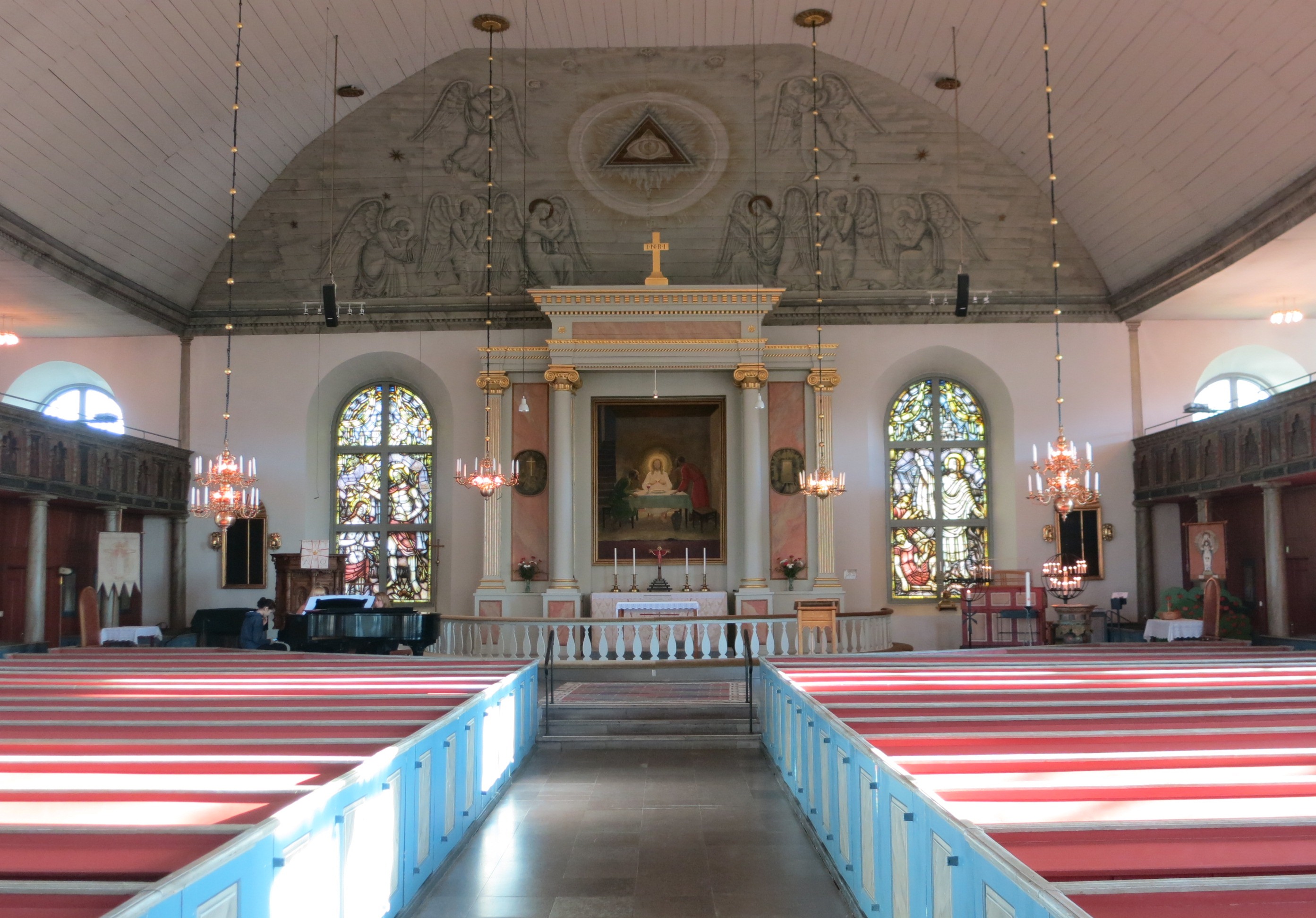 Osby kyrkas ljusa kyrkorum med bänkrader, ljuskronor och Olle Hjortzbergs Emmausmålning ovanför altaret.